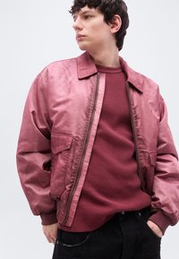 Νέος άνδρας με ξεθωριασμένο ροζ μπουφάν τύπου bomber πάνω από μια υφασμάτινη μπορντό μπλούζα, με τα χέρια στις τσέπες, κοιτάζοντας πλάγια μπροστά σε ένα απλό φόντο.