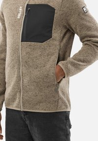 Veste en polaire marron clair avec une finition texturée. Dotée d'une poche poitrine noire, de poches latérales zippées et d'une coupe ajustée.