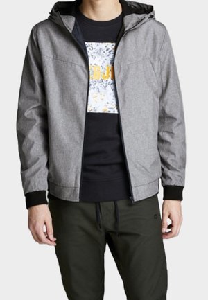 Uomo che indossa una giacca grigia con cappuccio e zip sopra una t-shirt nera con stampa grafica e pantaloni verde scuro con coulisse, in piedi con le mani rilassate lungo i lati.