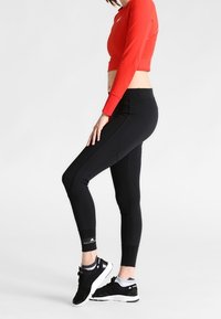 Donna in leggings neri e sneakers neri, solleva il piede sinistro, indossando un crop top rosso a maniche lunghe, mentre è in piedi su uno sfondo bianco.