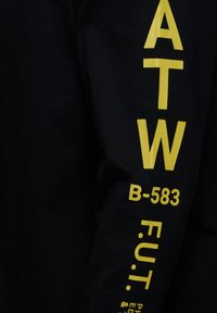 Svart långärmad tröja med djärv gul text på höger ärm, inklusive bokstäver och siffror. Tillverkad av bomull med en mjuk textur.