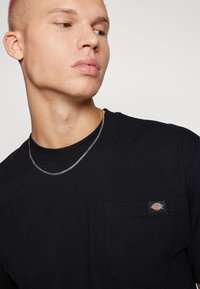 Maglietta nera a maniche lunghe con taschino, caratterizzata da una piccola patch con logo sul petto e scollo rotondo. Indossata con una collana in argento.