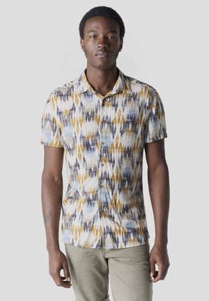 Joven con camisa de manga corta abotonada con patrones abstractos en azul, blanco y mostaza, y pantalones de color claro, de pie frente a un fondo gris.