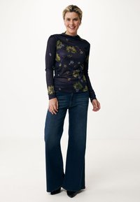 Långärmad marinblå topp med blommönster i grönt och lila, matchad med vida mörk denimjeans och svarta klackar. Slät textur och figursydd design.