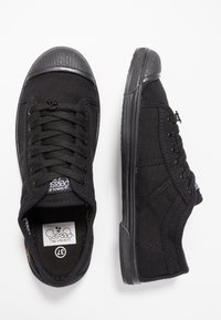 Sneaker in tela nera con punta in gomma, lacci neri piatti e suola testurizzata. Il lato presenta dettagli di cucitura e un piccolo etichetta logo.