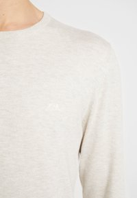 Pull beige clair à manches longues avec col rond. Présente un tricot texturé subtil et un petit logo de montagne brodé sur la poitrine.