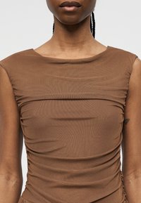 Buste supérieur d'une personne portant un haut marron sans manches froncé avec un col bateau, montrant une texture lisse et de légers plis de tissu.