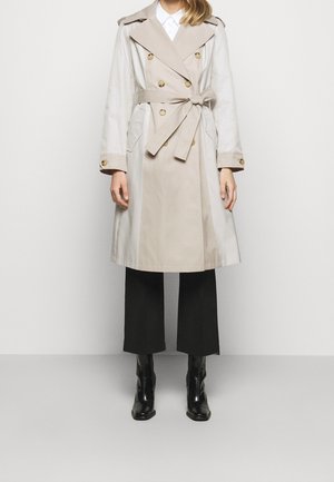 Femme portant un trench-coat beige et blanc à double boutonnage avec ceinture, pantalon noir court et bottines noires sur fond blanc.
