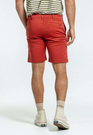 Shorts rouges en coton, dotés d'une poche arrière et d'un ourlet replié. Portés avec des baskets montantes beiges et des chaussettes beiges.