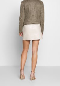 Chaqueta de ante verde oliva con un diseño ajustado, combinada con una minifalda brillante color crema. Tacones altos en rosa claro completan el conjunto.