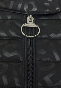 Sort pufferjakke med et blankt, prægede logo-mønster. Midterlukning med en cirkulær sølvhardware-detalje, der er mærket "GUESS."