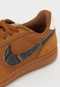 Sapatilha de camurça e couro castanho, com um swoosh da Nike em pele de cobra preta texturizada, atacadores castanhos e biqueira arredondada. Solado de borracha texturizada.