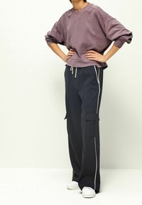 Nadměrná fialová mikina s kulatým výstřihem, ve spojení s tmavými kalhotami s bočními kapsami a bílým lemováním. Bílá teniska dokončuje vzhled.