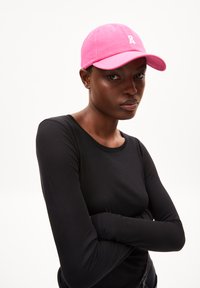 Rosa Baseballkappe mit gebogenem Schirm und weißen Logodetails, aus Baumwolle gefertigt, getragen mit einem langärmeligen schwarzen Oberteil vor einem weißen Hintergrund.