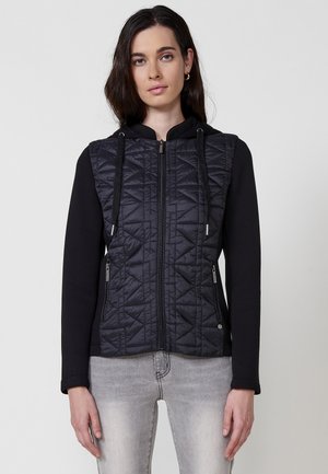 Chaqueta con cremallera que combina material acolchado negro y tela de jersey negra, con capucha, cordones ajustables y bolsillos laterales con cremallera.