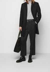 Cappotto nero su un dolcevita grigio, abbinato a pantaloni neri a gamba larga e scarpe nere. Tiene una borsa a spalla verde scuro. Texture liscia.