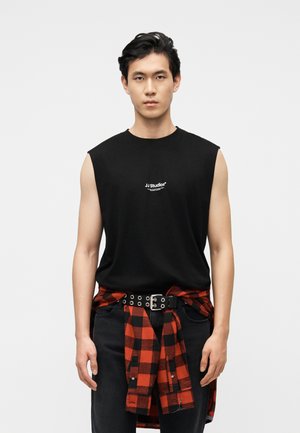 Άνδρας που φοράει μαύρο αμάνικο μπλουζάκι JJ Studios, μαύρο παντελόνι και κόκκινο-μαύρο καρό πουκάμισο δεμένο στη μέση, σε απλό φόντο.