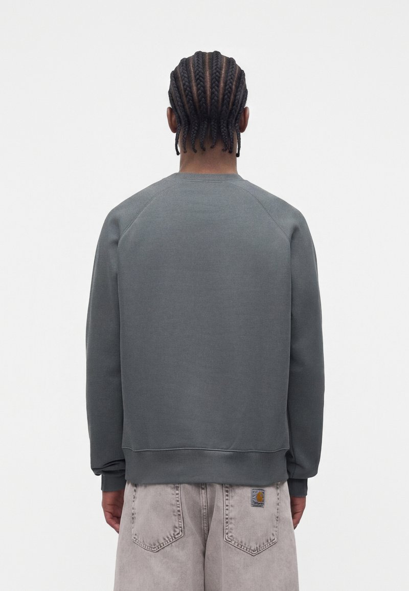Person mit Cornrow-Zöpfen trägt einen dunkelgrauen Sweatshirt und hellgraue Jeans, steht mit dem Rücken zur Kamera vor einem schlichten weißen Hintergrund.