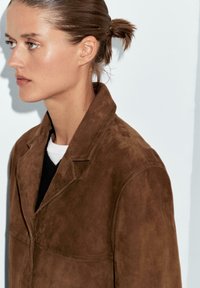 Bruine suède jacket met een notch-kraag, voorzakken en zichtbare stiksels, gedragen over een zwarte trui.