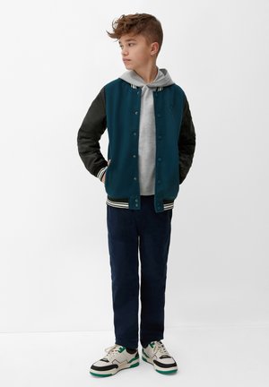 Veste varsity en laine vert émeraude avec des manches en cuir noir, col rayé, un sweat à capuche gris superposé en dessous, associée à un pantalon bleu marine et des baskets blanches.