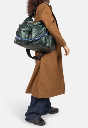 Persoon draagt een bruine trenchcoat en donkere spijkerbroek en houdt een grote glanzende groene totebag vast met een blauw gevlochten handvatdetail.