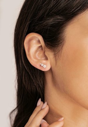 Silberne Steckohrringe mit floralem Design, verziert mit kleinen klaren Edelsteinen, getragen am Ohr eines Models. Im Hintergrund sind dunkles Haar und Haut zu sehen.