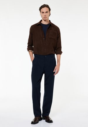 Uomo in piedi con le mani in tasca, indossa una camicia marrone scuro abbottonata, pantaloni blu navy e scarpe marrone scuro su sfondo bianco.