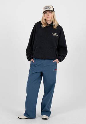 Persona con una sudadera negra de gran tamaño con el texto "Prohibido", pantalones anchos azules, una gorra beige y negra, y zapatos blancos, de pie con las manos en los bolsillos.