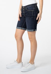 Donkerblauwe denimshorts met opgerolde zomen, voorzien van een vijf-knopen ritssluiting, voorkruiszakken en decoratieve stiksels aan de zijkanten.