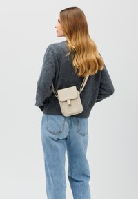 Borsa a tracolla beige con chiusura a patta, forma rettangolare, texture liscia e tracolla regolabile, indossata su un maglione grigio e jeans blu.