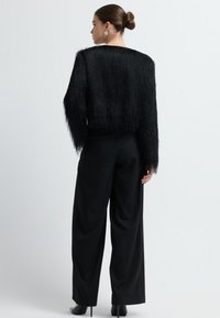 Kobieta z włosami w koku, ubrana w czarną, teksturowaną bluzkę z długim rękawem, szerokie czarne spodnie i czarne szpilki, odwrócona plecami.