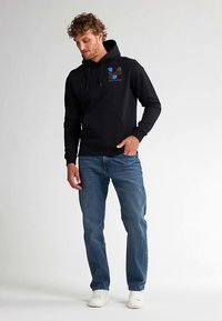 Felpa nera con cappuccio con coulisse, tasca frontale e design grafico multicolore sul petto a sinistra. Abbinata a jeans blu e sneakers bianche.
