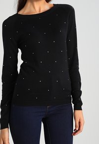 Svart stickad tröja med långa ärmar och lös passform, prydd med gyllene prickar som är jämnt fördelade över framsidan.