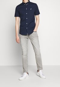 Uomo che indossa una camicia button-up a maniche corte fitted di colore blu navy, jeans slim grigi chiari e sneaker bianche, in posa con una mano in tasca.