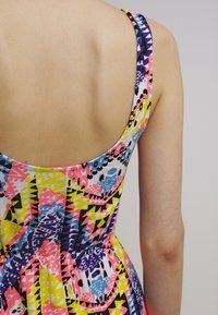 Le dos et le bras d'une personne portant une robe sans manches colorée avec des motifs géométriques et abstraits en rose, jaune, violet, bleu et noir.