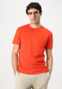 Camiseta de algodón roja con cuello redondo, mangas cortas y un pequeño detalle del logo en el pecho, combinada con pantalones cortos de color beige claro.