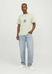 Vīrietis stāv ar roku kabatā, valkājot gaiši zaļu T-kreklu ar logo, brīvus gaiši zilos džinsus un baltas sporta kurpes pret vienkāršu fona.