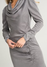Abito grigio con scollatura a barca, maniche lunghe e dettagli arricciati sui fianchi. Tessuto morbido e testurizzato con un leggero luccichio e una silhouette aderente.