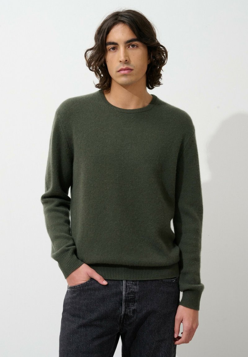 Maglione a maglia verde scuro con collo rotondo e polsini a coste. Il tessuto appare morbido, offrendo una vestibilità comoda. Indossato con jeans grigio scuro.