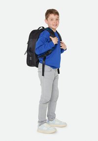 Zaino in tessuto nero con spallacci regolabili, indossato da un bambino in una felpa blu, jeans grigi e scarpe azzurro chiaro. Design semplice.