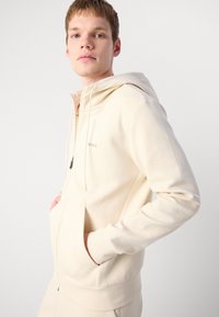 Cremefarvet hættetrøje med lynlås, justerbar snor til hætten og sidelommer. Glat stof og minimalistisk branding på brystet.