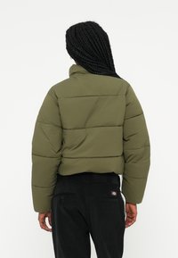 Giacca a vento verde oliva con design cropped, motivo trapuntato e colletto alto. Indossata sopra pantaloni neri con tasche posteriori.