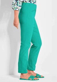 Grüne Baumwollhosen im Slim-Fit mit einem Reißverschluss-Rücksack, kombiniert mit passenden grünen Sandalen mit einem Ausschnitt-Design.