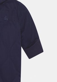 Marineblauwe gewatteerde jas met korte mouwen, voorzien van een ribgebreide manchette en een subtiele tonale logo op de borst. Gladde textuur met een modern ontwerp.