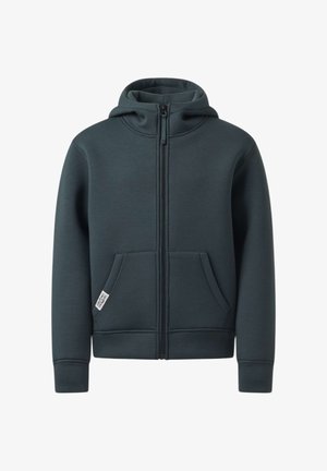 Garçon debout, main sur la nuque, portant un sweat à capuche zippé foncé, un jean bleu clair et des baskets blanches avec des rayures noires.