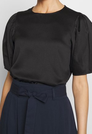 Femme portant un chemisier noir à manches bouffantes rentré dans un pantalon taille haute bleu marine avec une ceinture en tissu nouée en nœud.