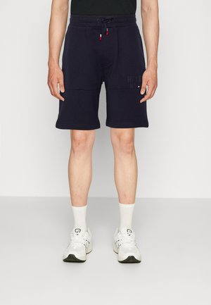 Persona vistiendo pantalones cortos con cordón azul marino, calcetines blancos hasta la pantorrilla y zapatillas blancas sobre un fondo claro y sencillo.