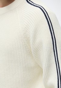 Pull blanc en maille avec texture côtelée et deux bandes noires parallèles courant le long de l’épaule et de la manche.