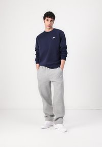 Donkerblauwe sweatshirt met een klein wit logo, gecombineerd met lichtgrijze trainingsbroeken. Model staat tegen een egale achtergrond en draagt witte sneakers.