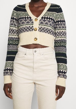 Personne à la peau foncée portant un cardigan court en maille crème avec des rayures à motifs et de gros boutons, associé à un pantalon en velours côtelé crème taille haute.